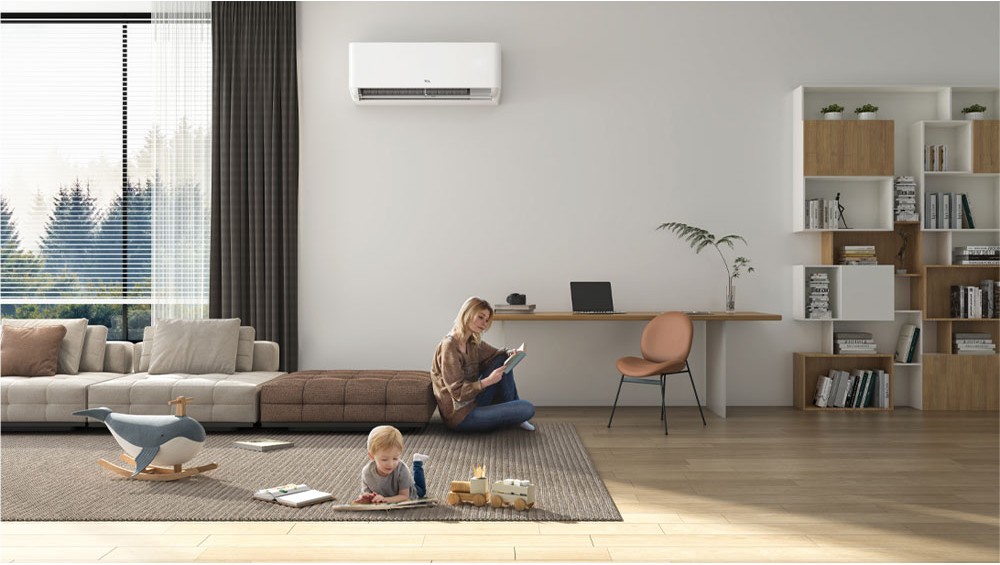 A woman reads a book while sitting on a brown ottoman in a living room, with a young child playing on the rug in front of her. A white TCL air conditioner is mounted on the wall above her.